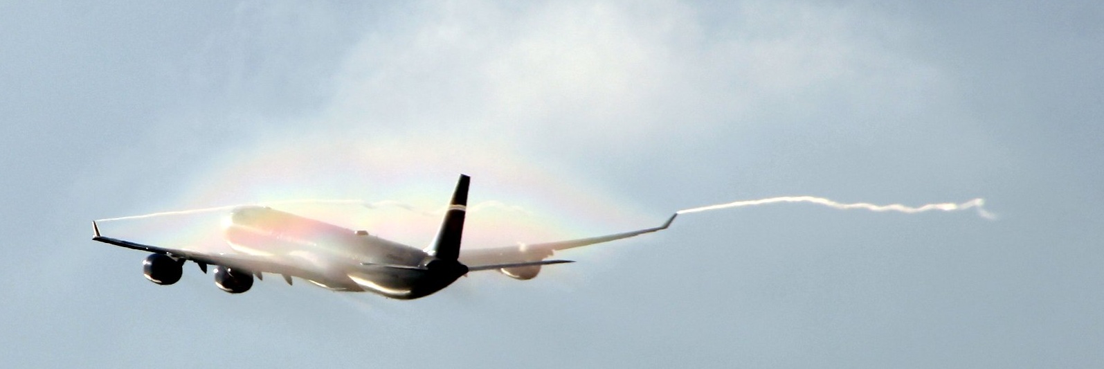 A large airplane departing into  blue skes with contrails coming off the wingtips and a rainbow forming in the moisture above it